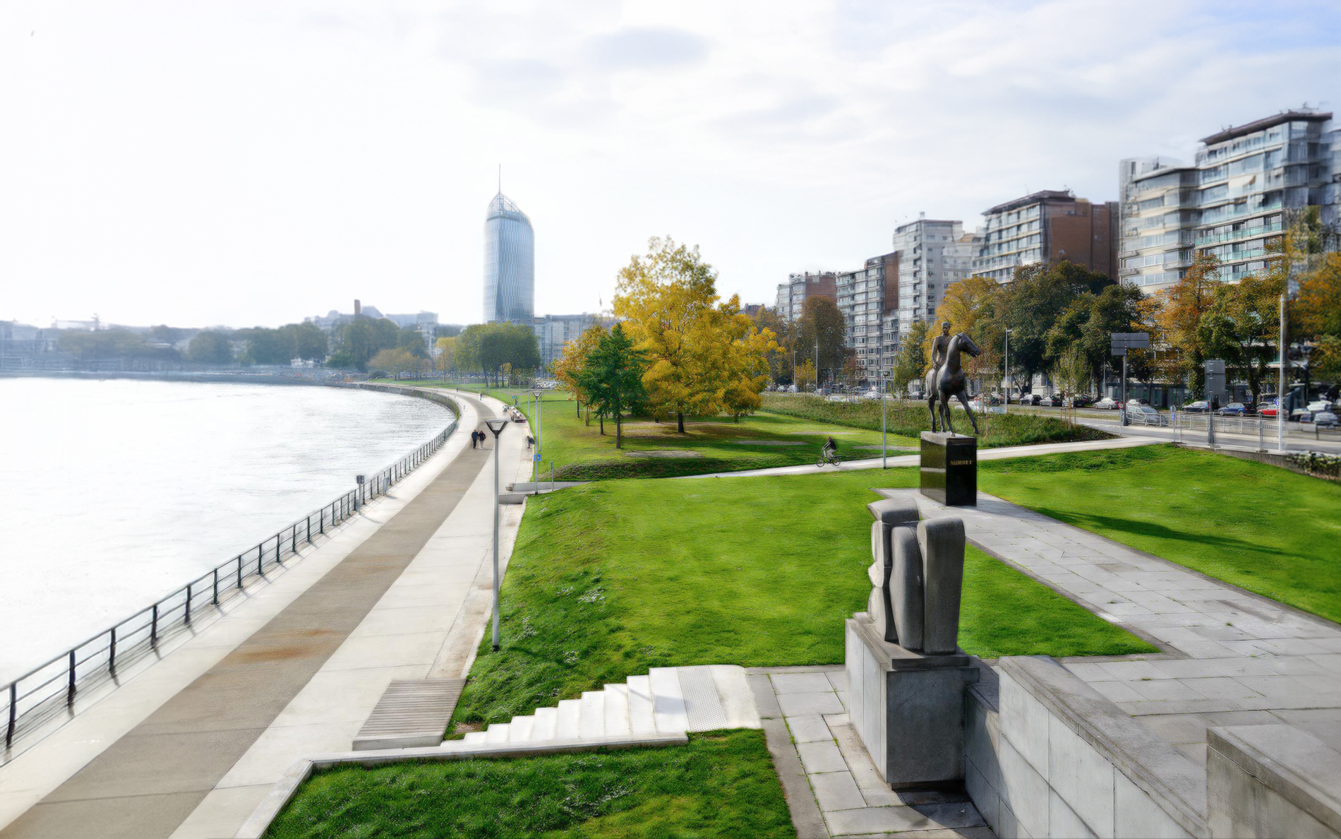 Quai le long de la Meuse à Liège avec un chemin piétonnier, des pelouses vertes, des arbres aux feuilles d'automne, une statue équestre, et des immeubles en arrière-plan.